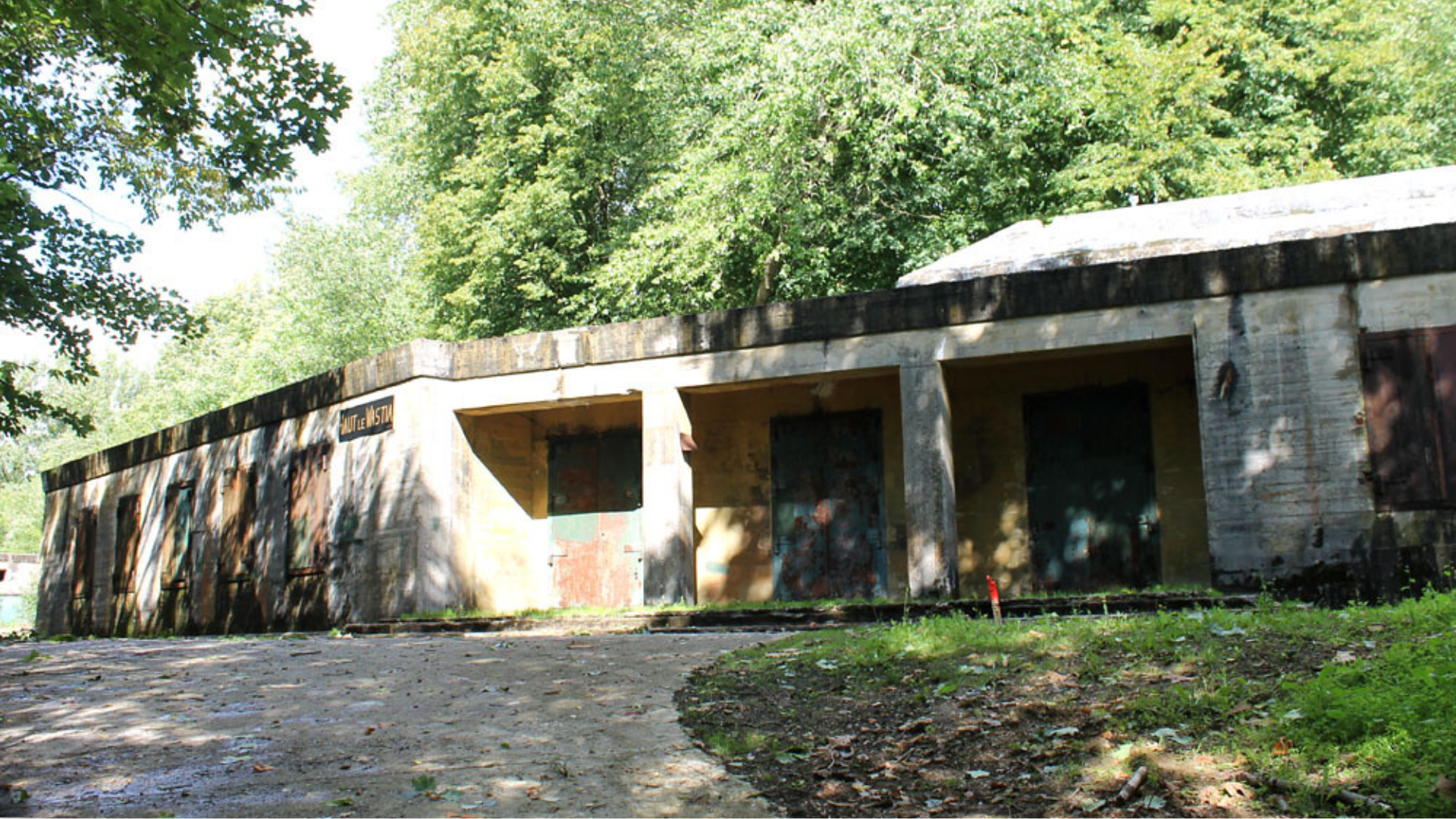 Ancien bâtiment en béton avec une série de portes et de colonnes, situé au bord d’un chemin dans une zone boisée ; une plaque au mur indique “Haut le Wastia”.